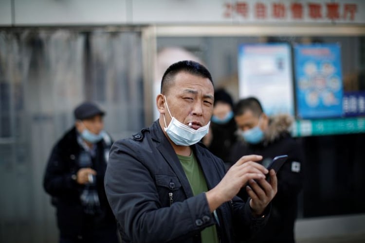 Un hombre con una máscara facial usa su teléfono móvil mientras fuma fuera de la estación de ferrocarril de Beijing, China (REUTERS)