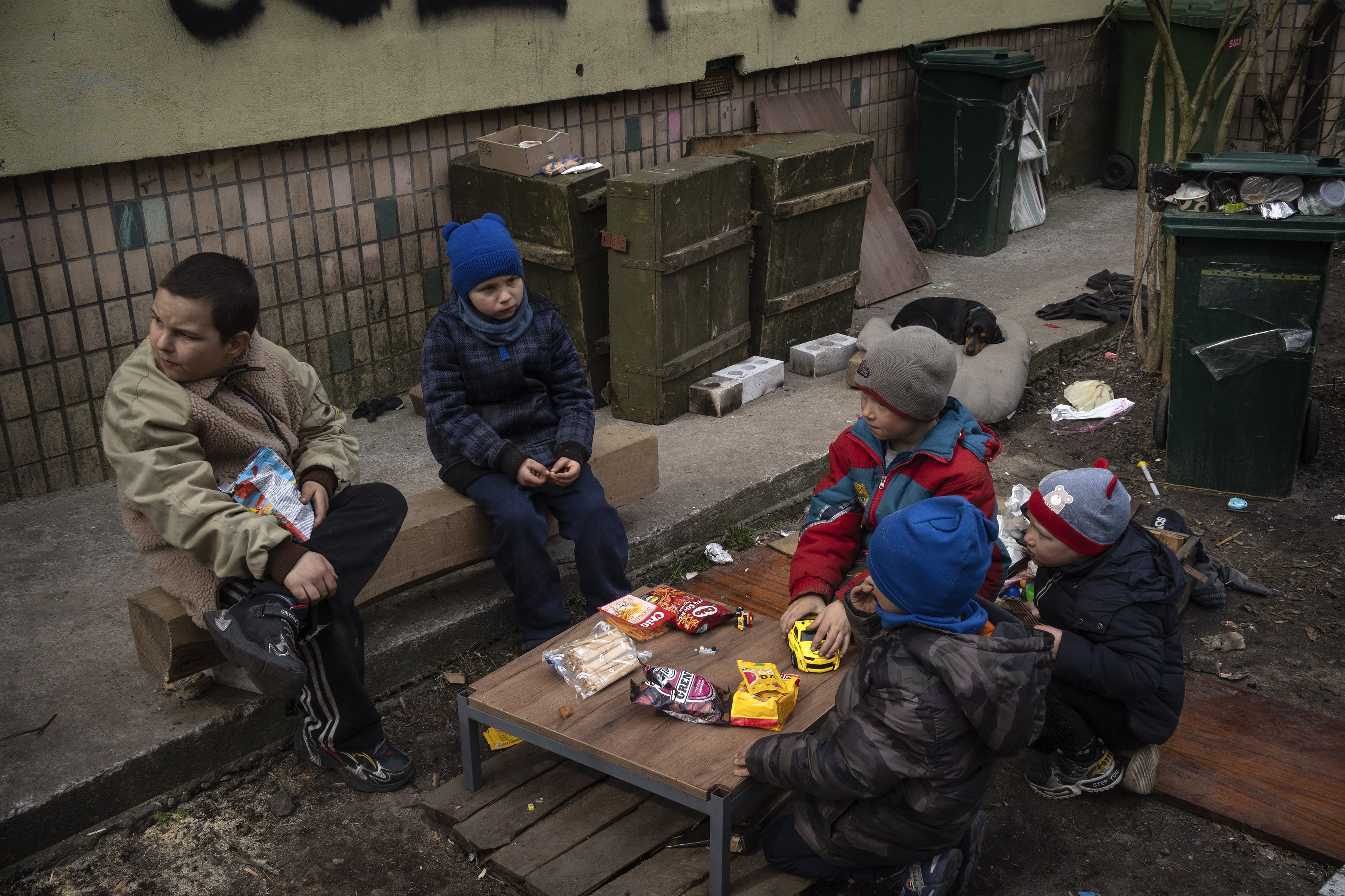 Niños sentados en torno a una mesa pequeña con comida que recibieron en un reparto de comida gratuita en Bucha, a las afueras de Kiev, Ucrania, el viernes 8 de abril de 2022. Un graffiti escrito por los vecinos en la pared al fondo dice "Niños" para informar al ejército ruso de que muchos niños viven en el edificio. (AP Foto/Rodrigo Abd)