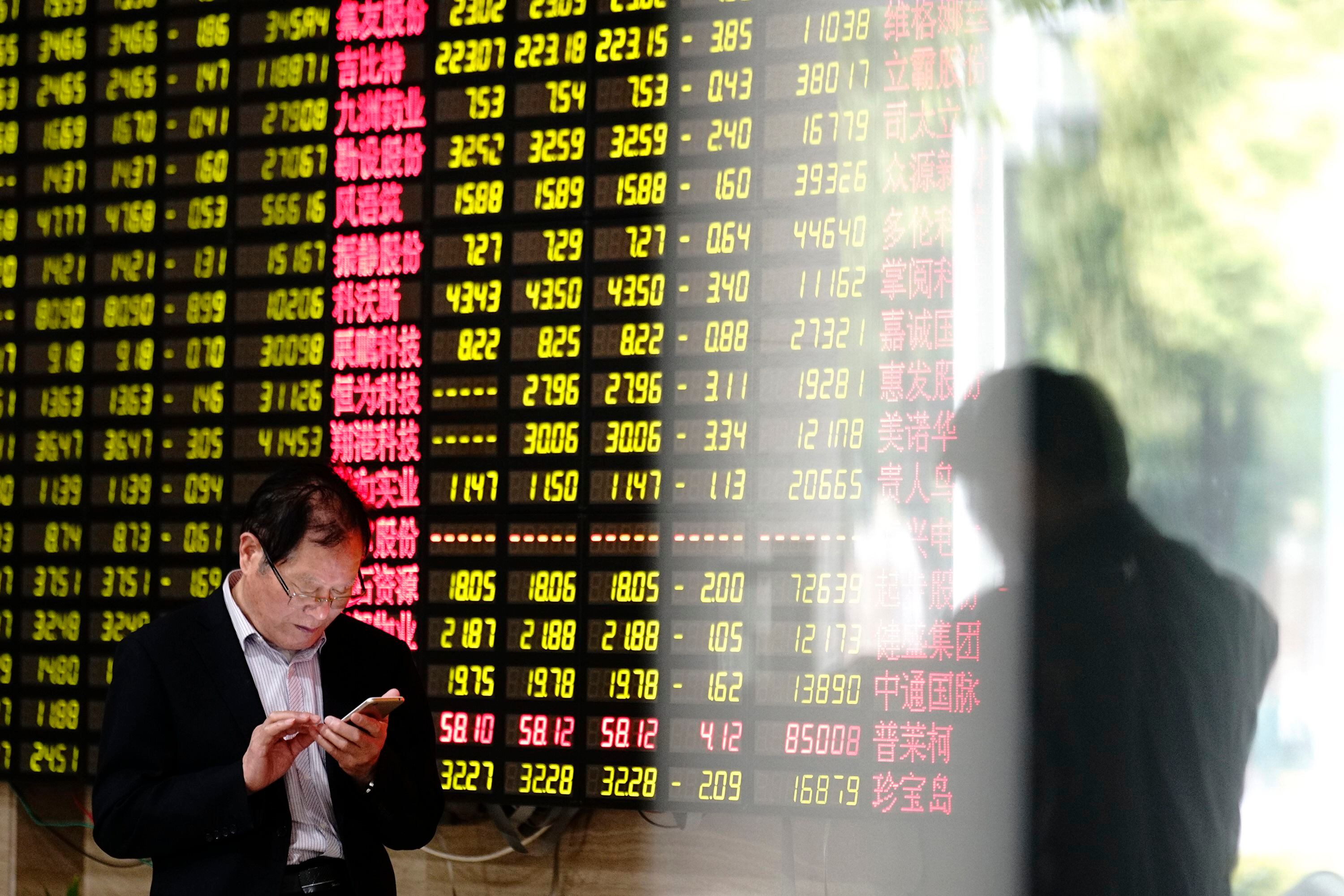 Un hombre mira su teléfono inteligente cerca de una pantalla que muestra los precios de las acciones en una casa de bolsa en Shanghai. (Foto AP)