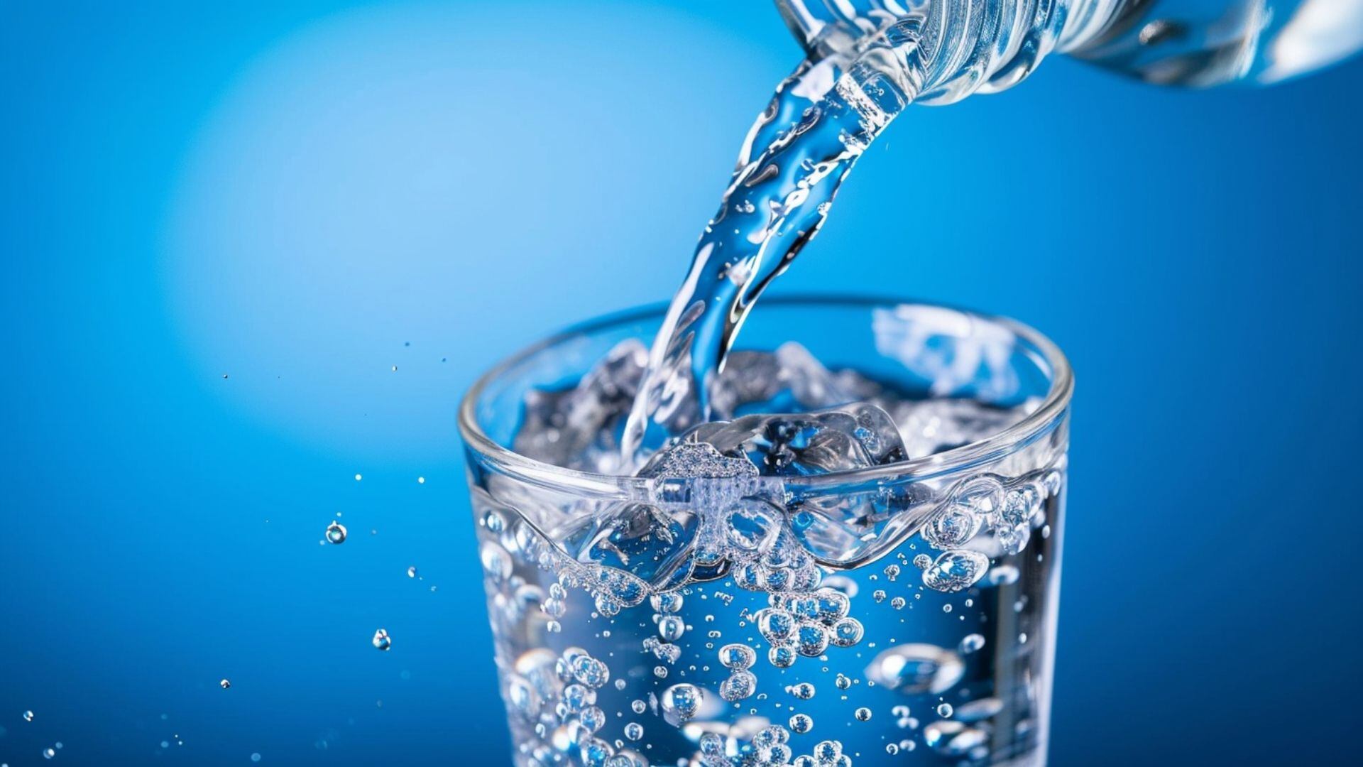 Una botella transparente de agua sirve líquido sobre un vaso de vidrio, burbujas, splashes,, fondo azul, refrescante, sed, publicidad de bebida, tomar, beber, consumir líquido, H2O - visualesIA