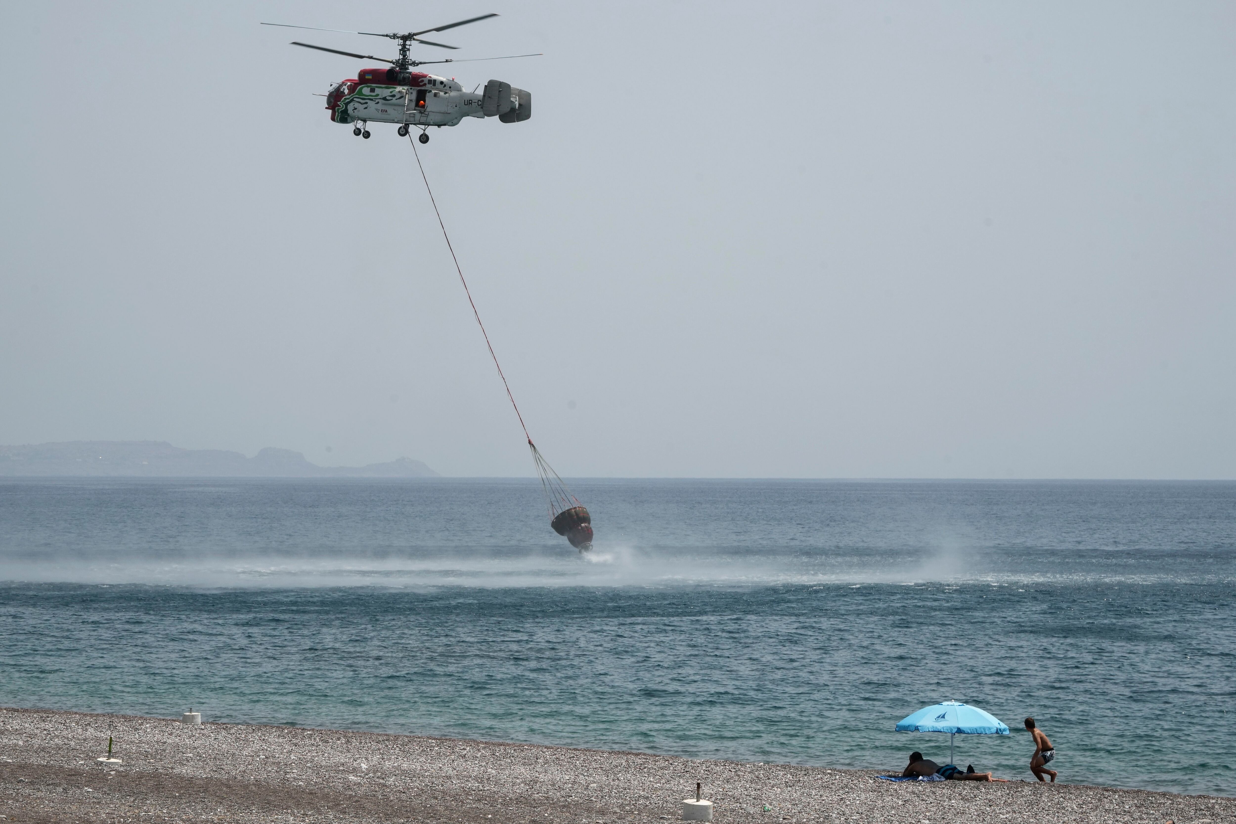Dos personas en la playa observan como un helicóptero carga una cesta de agua en el mar en un operativo de extinción de incendios. (AP Foto/Petros Giannakouris)