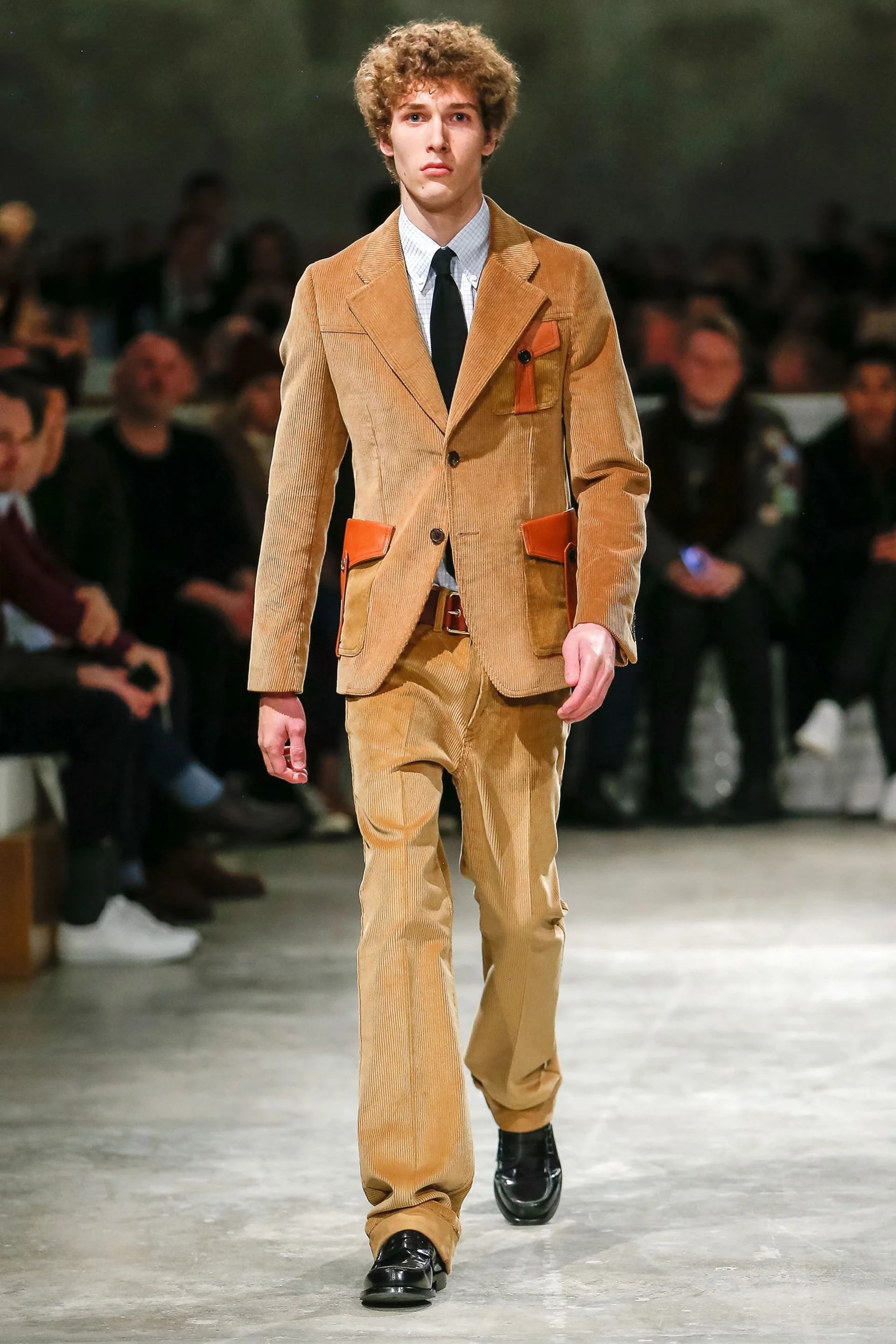 Prada, la casa italiana, lució en pasarela un traje camel con detalles de solapas de bolsillos en naranja acompañado un look formal con camisa blanca y corbata.