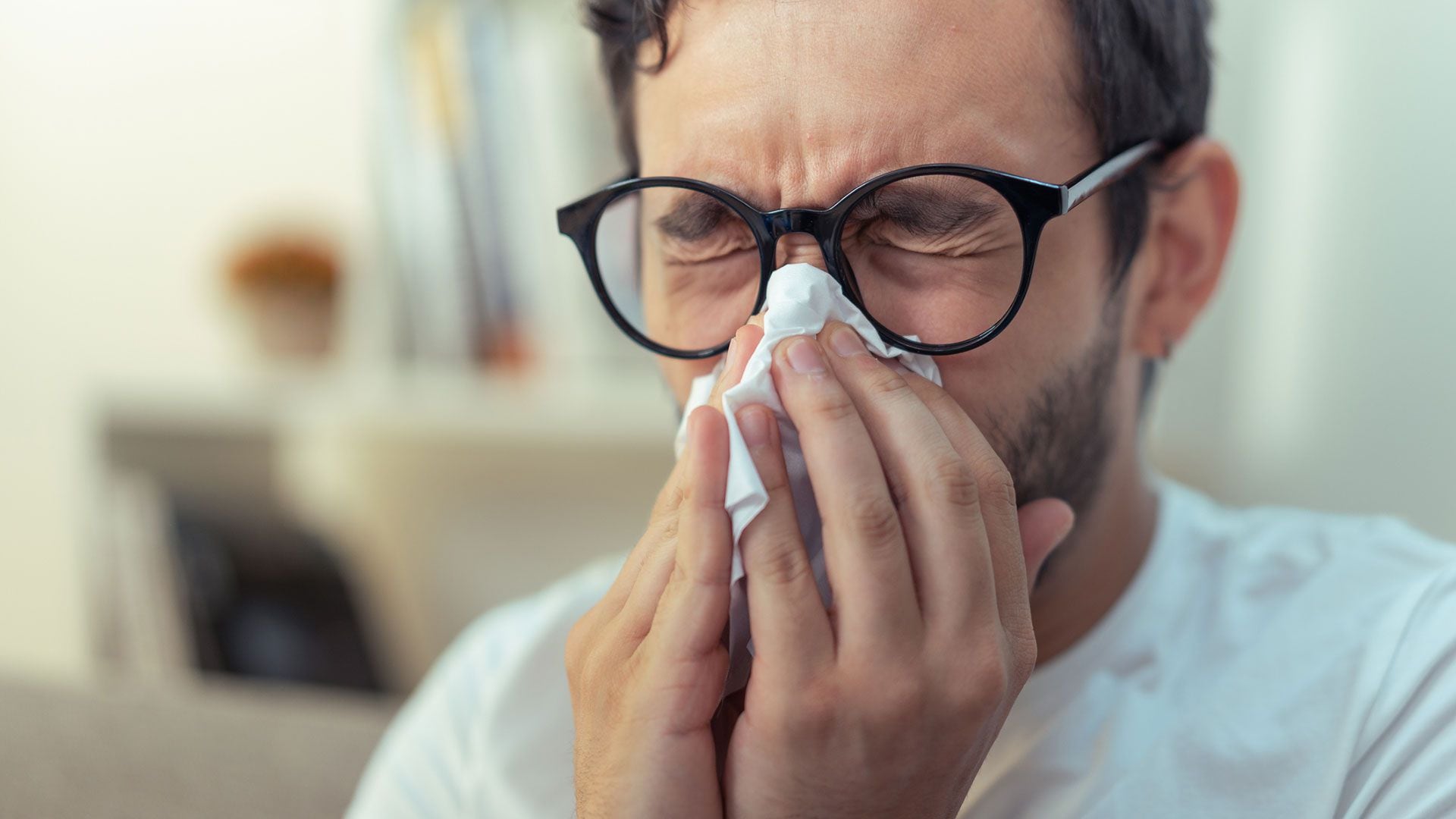 Young man with glasses sneezing, wiping his nose with a piece of tissue paper. (Getty)