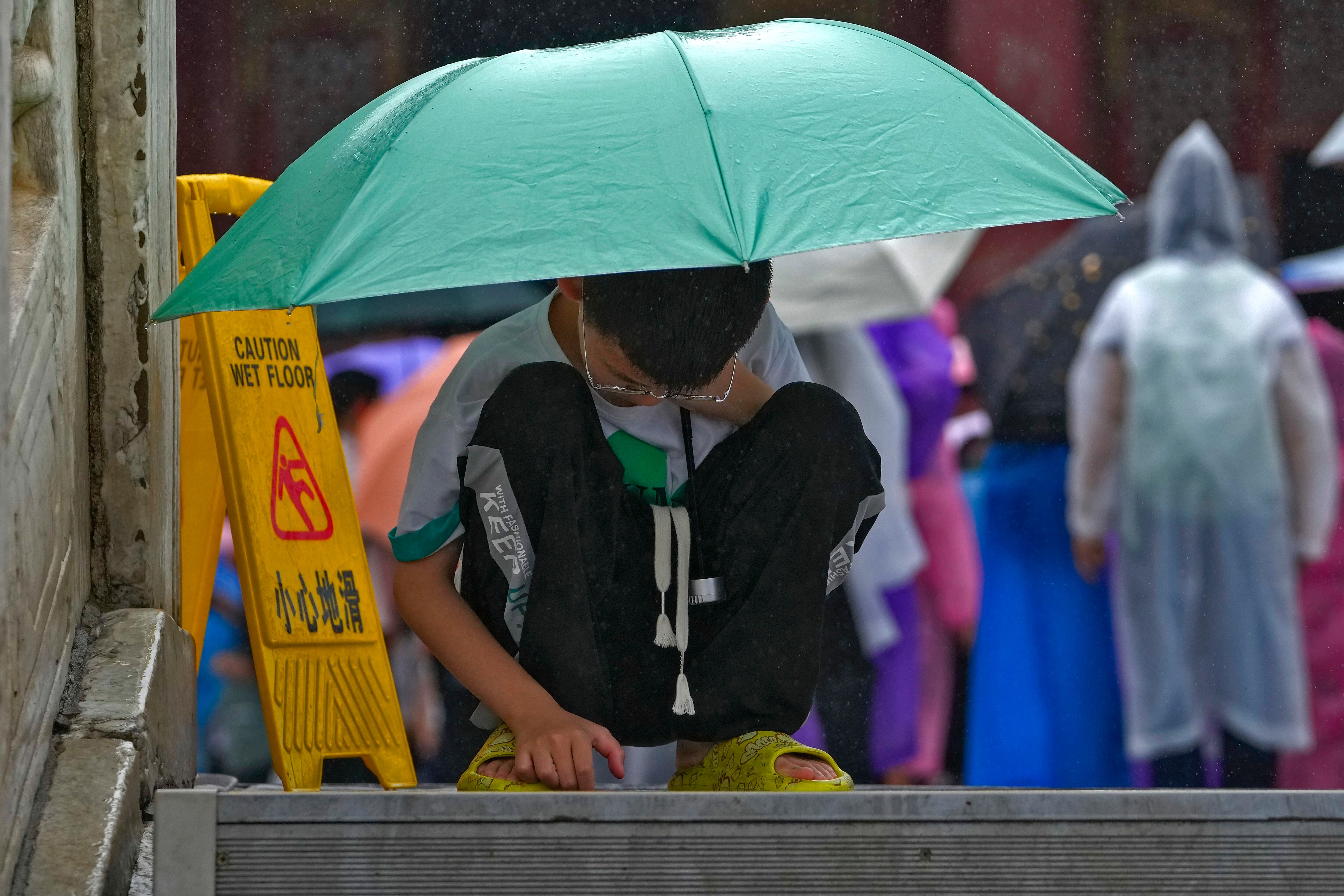 Un niño se protege de la lluvia con un paraguas mientras revisa un celular, en el piso de la Ciudad Prohibida de Beijing (AP Foto/Andy Wong)