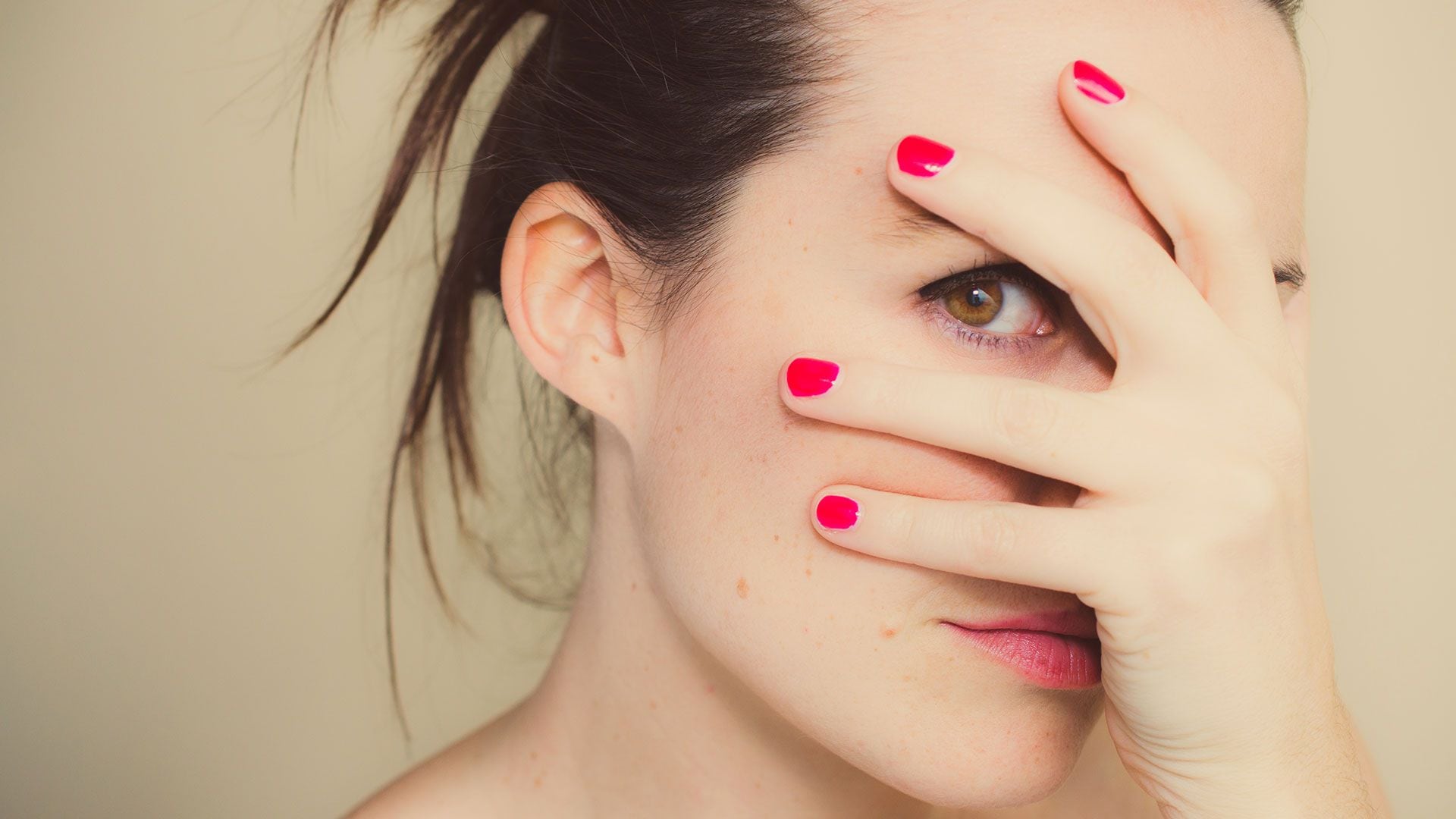 Close-up portrait. Girl covering her face with one hand and looking between her fingers. One brown eye and gathered hair. (Getty)
