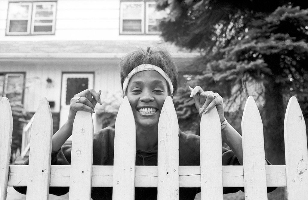 La cantante y actriz estadounidense Whitney Houston mira por encima del portón de la casa de su madre, West Orange, Nueva Jersey, el 28 de mayo de 1985. (Photo by Jack Vartoogian/Getty Images)