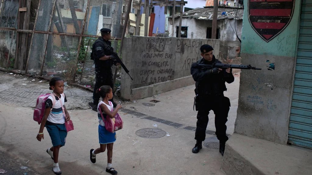 Policías apuntan con armas largas mientras dos niñas van a la escuela caminando como si nada pasara en la favela Complexo do Alemao (AP)