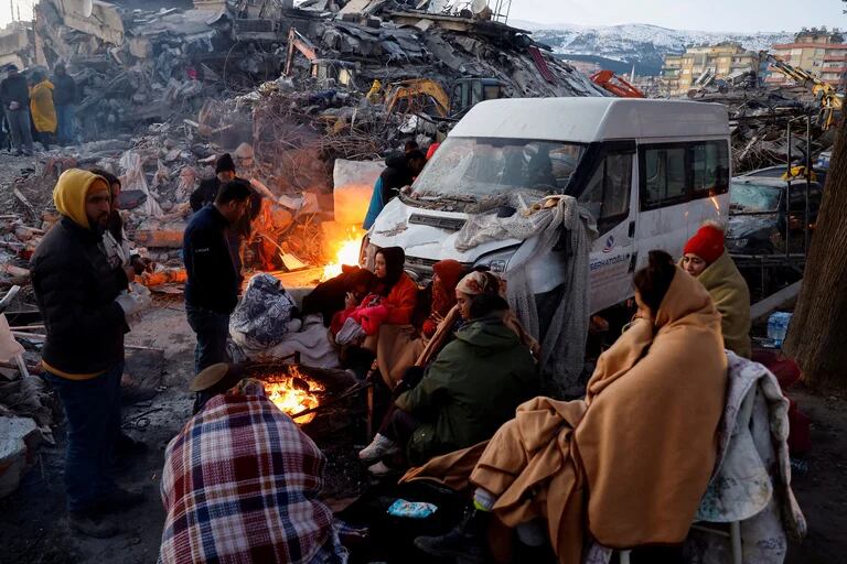 La gente se sienta alrededor de un fuego junto a los escombros y los daños cerca del sitio de un edificio derrumbado en  