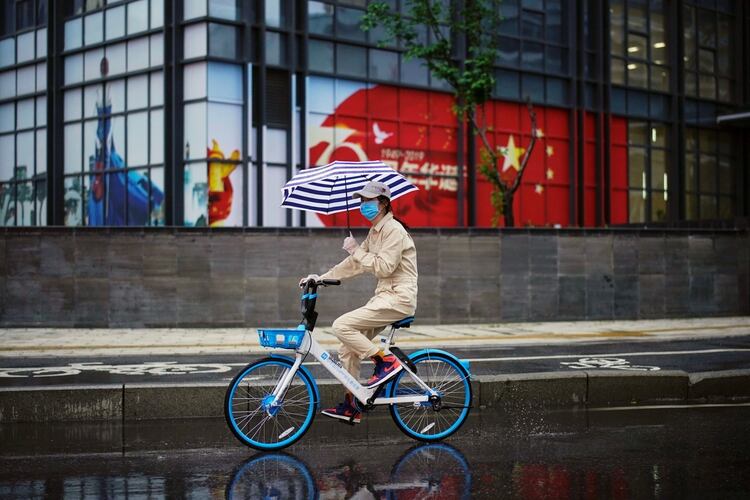 Una mujer con un paraguas monta una bicicleta compartida junto a una imagen de la bandera china después de que se levantó la cuarentena en Wuhan, la capital de la provincia de Hubei y el epicentro del brote de (Reuters)