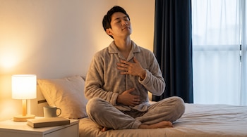 Hombre joven sentado en una cama con pijama, manos sobre el pecho y abdomen, ojos cerrados. Una lámpara, libro y taza en la mesa de noche.