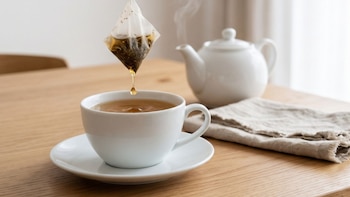 Una taza de cerámica sobre un plato, una bolsa filtrante suspendida que gotea, una tetera y una servilleta en una mesa de madera