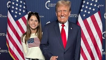 Donald Trump con traje azul y corbata roja sonríe junto a Kai Trump frente a dos banderas de Estados Unidos y un fondo azul