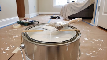 Una lata de pintura blanca con una banda elástica sostiene una brocha que gotea en una habitación con papel protector en el suelo.