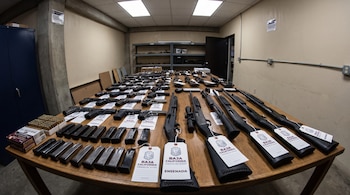 Mesa de madera con pistolas, escopetas, rifles, cargadores y cartuchos apilados. Se observan etiquetas del Gobierno de Baja California.