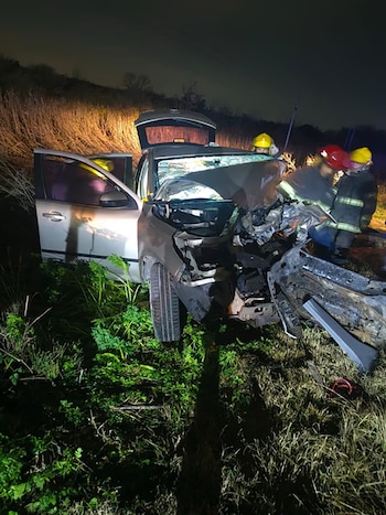 Automóvil plateado fuertemente dañado con puertas abiertas sobre pastos. Tres bomberos con uniformes y cascos trabajan en la escena nocturna