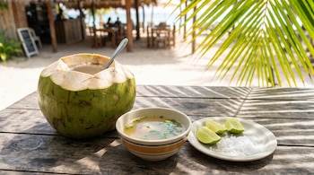 Agua de coco en un coco verde, un tazón de consomé, y un plato con limón en rodajas y sal, sobre una mesa de madera. Palmeras y playa al fondo.