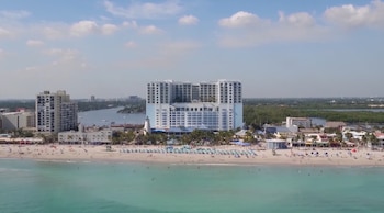 Vista aérea de una playa en Florida con un gran resort blanco y azul, personas en la arena y el agua turquesa del océano, y un canal detrás