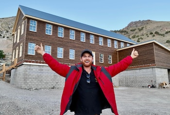 Hombre con chaqueta roja y gorra negra levanta los brazos frente a un edificio grande de madera con base de bloques de concreto