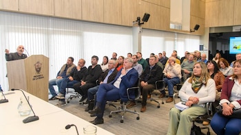 Un orador habla desde un atril con el escudo de La Pampa ante un grupo de personas sentadas en una sala de reuniones