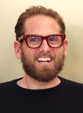 Primer plano del actor Jonah Hill sonriendo, con cabello castaño, barba y gafas de montura roja, sobre un fondo liso de color beige