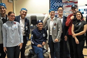 Un grupo de personas posa de pie y un hombre permanece sentado frente a una consola médica blanca ante un cartel de AdventHealth