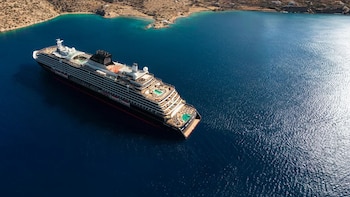 Vista aérea de un gran crucero, el Explora I, navegando en un mar azul intenso cerca de una costa rocosa y árida con playas de arena