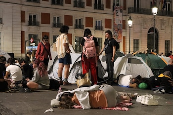 Varias personas conversan y descansan en el suelo junto a tiendas de campaña frente a un edificio iluminado de noche