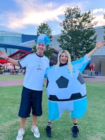 Un hombre y una mujer posan sonrientes en un campo de césped. El hombre lleva sombrero de Argentina, camiseta blanca y shorts. La mujer lleva un disfraz inflable de balón de fútbol