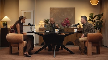 Michelle Obama, Jimmy Kimmel y otro hombre ríen y conversan alrededor de una mesa oscura con micrófonos en un estudio de podcast, rodeados de decoración