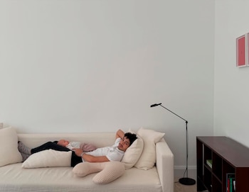 Chino Darín recostado en un sofá blanco con un bebé dormido a su lado. El hombre sonríe a la cámara, apoyado en almohadas, en una sala con paredes blancas