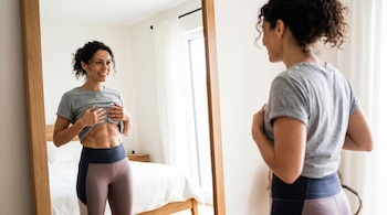 Mujer de unos 40 años con ropa deportiva, levantando su camiseta para mostrar abdominales tonificados frente a un espejo de cuerpo entero en un dormitorio.