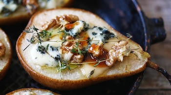 Primer plano lateral de media pera con queso azul fundido, nueces y tomillo, mostrando la pulpa y la textura del queso, sobre fondo oscuro y desenfocado.