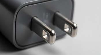 Primer plano de las dos clavijas metálicas de un cargador de celular de color gris oscuro, sobre un fondo blanco, con la clavija frontal en foco.