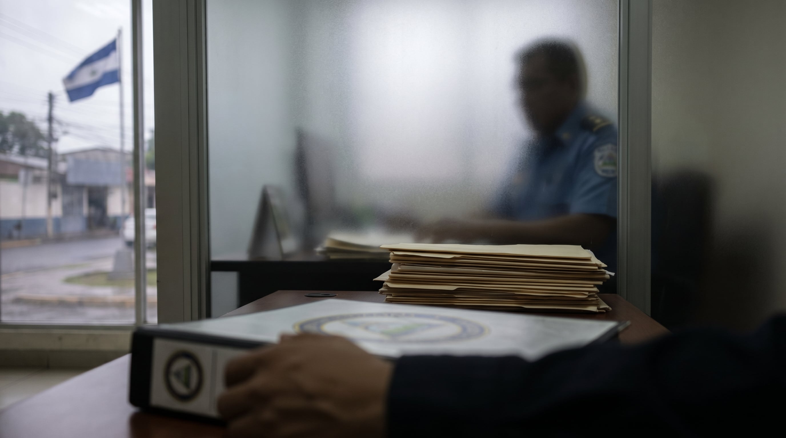 Una mano sostiene un documento mientras una pila de expedientes se observa en una oficina donde, tras un cristal esmerilado, una figura se mantiene oculta, reflejando la opacidad estatal del MINSA en Nicaragua. (Imagen Ilustrativa Infobae)