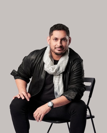 Retrato de Rodrigo Parra, un hombre con chaqueta de cuero negra y bufanda estampada, sentado en una silla negra mirando al frente