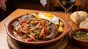 Un plato de bife a la criolla en una cazuela de barro, con carne, salsa de tomate y verduras, un huevo frito, patatas fritas, pan y vino tinto.