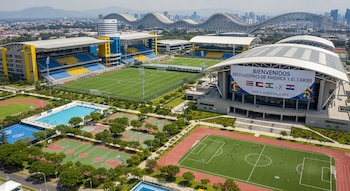 Vista aérea de complejo deportivo en Ciudad de Guatemala. Estadios, campos, pista, canchas, piscina. Cartel 'BIENVENIDOS' visible. Ciudad al fondo.