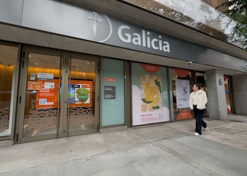 Una mujer de abrigo claro y pantalones oscuros camina sobre la vereda frente a la sucursal de un Banco Galicia. La fachada tiene ventanales con rótulos y pantallas