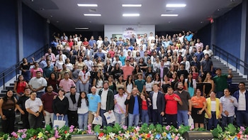 Una multitud de personas jóvenes y adultas de diversas etnias posan para una foto grupal en un auditorio. Hay escaleras a los lados y decoraciones florales al frente