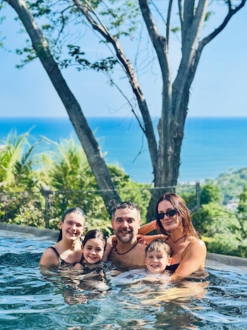 Una mujer, un hombre y tres niños se encuentran en una piscina con el mar, árboles y vegetación de fondo en un día soleado