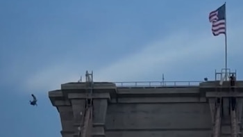 Un hombre cae junto a la estructura de hormigón de un puente. Una bandera de Estados Unidos ondea en un mástil sobre el puente. Cielo azul