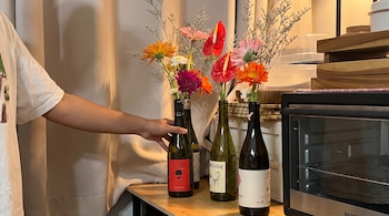 Una persona sostiene una botella de vidrio con flores junto a otras botellas y un horno sobre un mueble de madera.