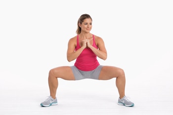 Una mujer con cabello recogido realiza una sentadilla, vista de frente, viste top rosa, pantalones cortos grises y zapatillas deportivas.