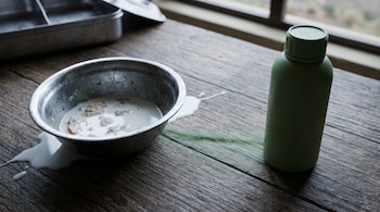 Un cuenco metálico con leche y restos de comida sobre una mesa de madera, junto a una botella verde y una bandeja metálica al fondo.