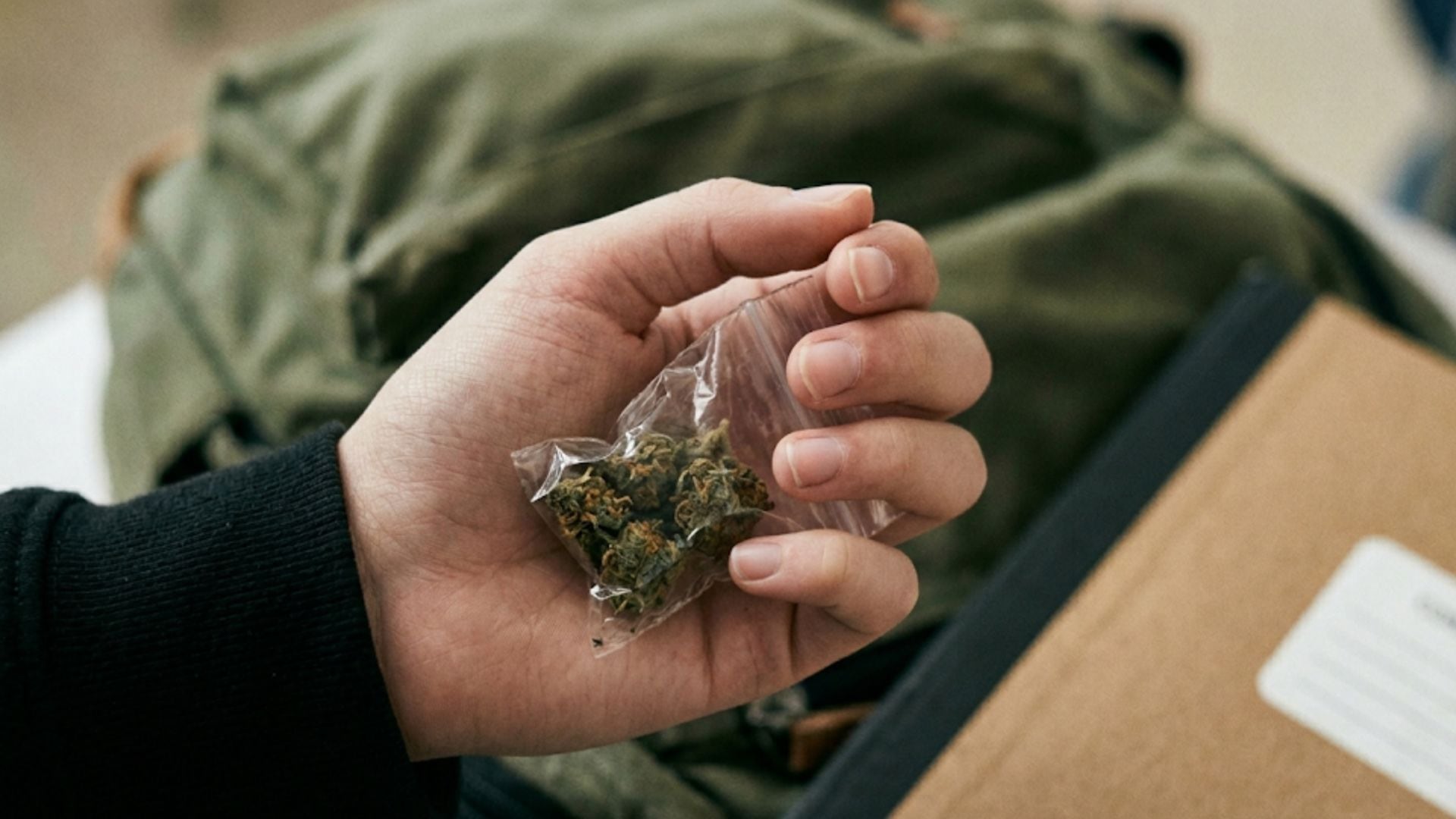 Una mano adolescente sostiene discretamente una pequeña bolsa con cogollos de cannabis, con el fondo desenfocado de una mochila y un cuaderno escolar. (Imagen Ilustrativa Infobae)