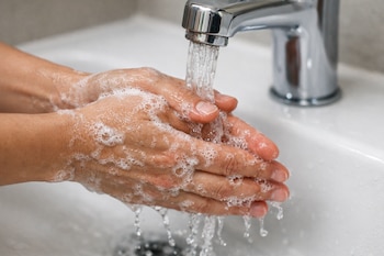 Manos de persona lavándose con jabón bajo el agua de un grifo metálico sobre un lavamanos blanco.