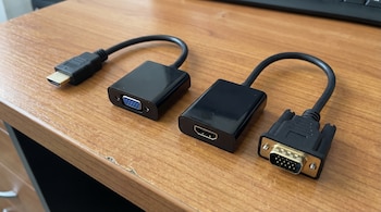 Dos adaptadores de video negros sobre un escritorio de madera. Uno tiene un conector HDMI macho y un puerto VGA hembra, el otro tiene HDMI hembra y VGA macho.