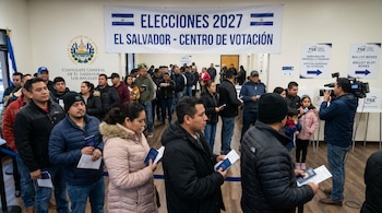 Larga fila de personas dentro de un consulado, esperando para votar. Un cartel dice "Elecciones 2027 El Salvador - Centro de Votación". Periodistas filman.