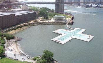 Vista aérea de una piscina flotante en forma de cruz con personas nadando en el East River de Nueva York, cerca de la orilla y el Puente de Brooklyn