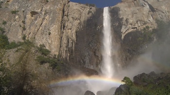 Gran cascada blanca cae por una pared de roca gris y marrón. Hay niebla en la base y un arcoíris de colores brillantes justo encima del agua. Se ven árboles